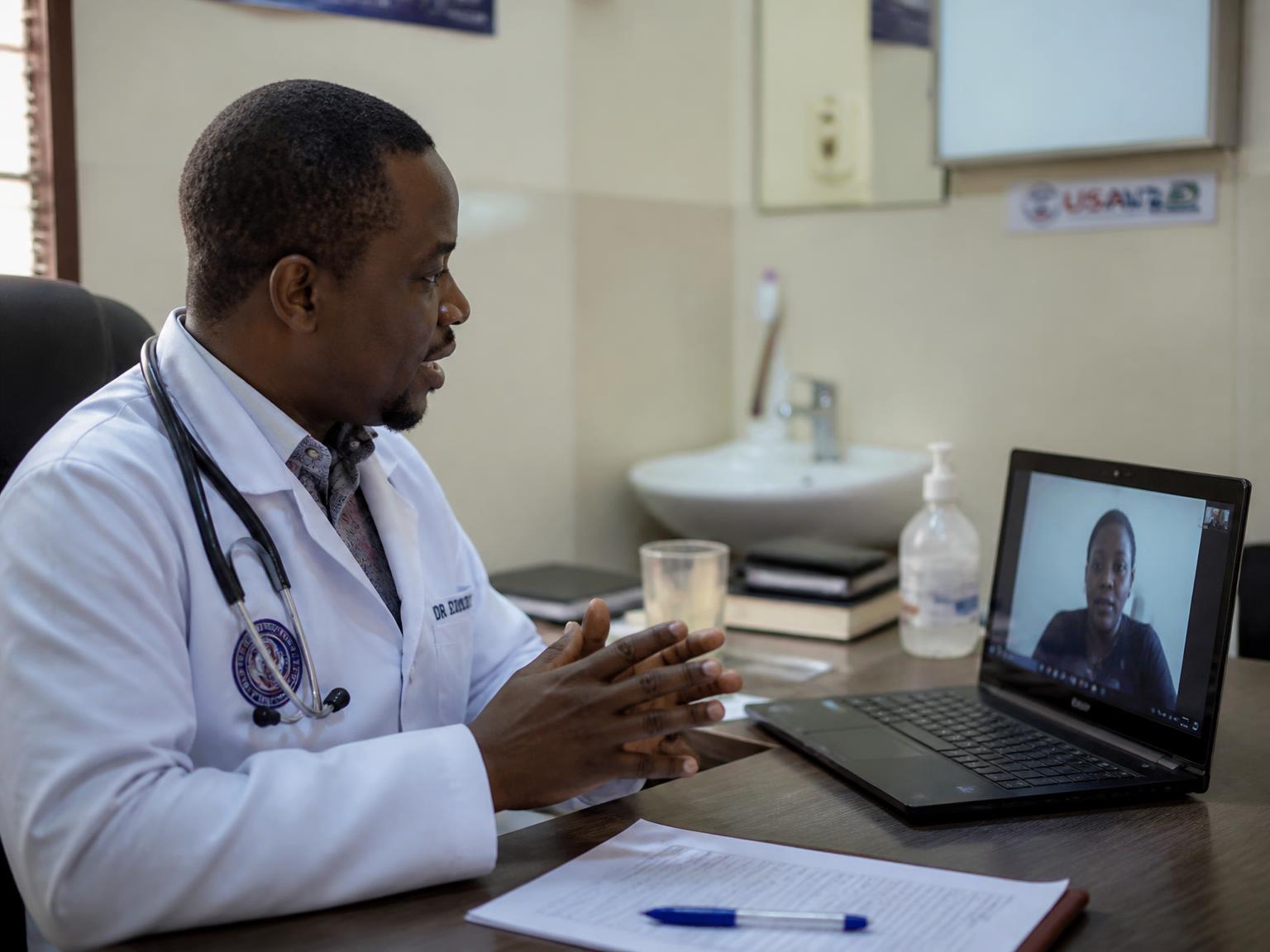 A doctor in a white coat speaking with a patient over a video call on a laptop