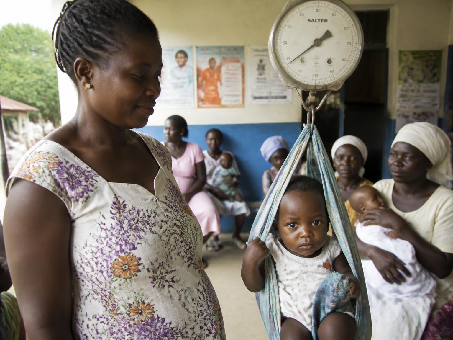 A toddler being weighed on a hanging scale at a community child welfare clinic