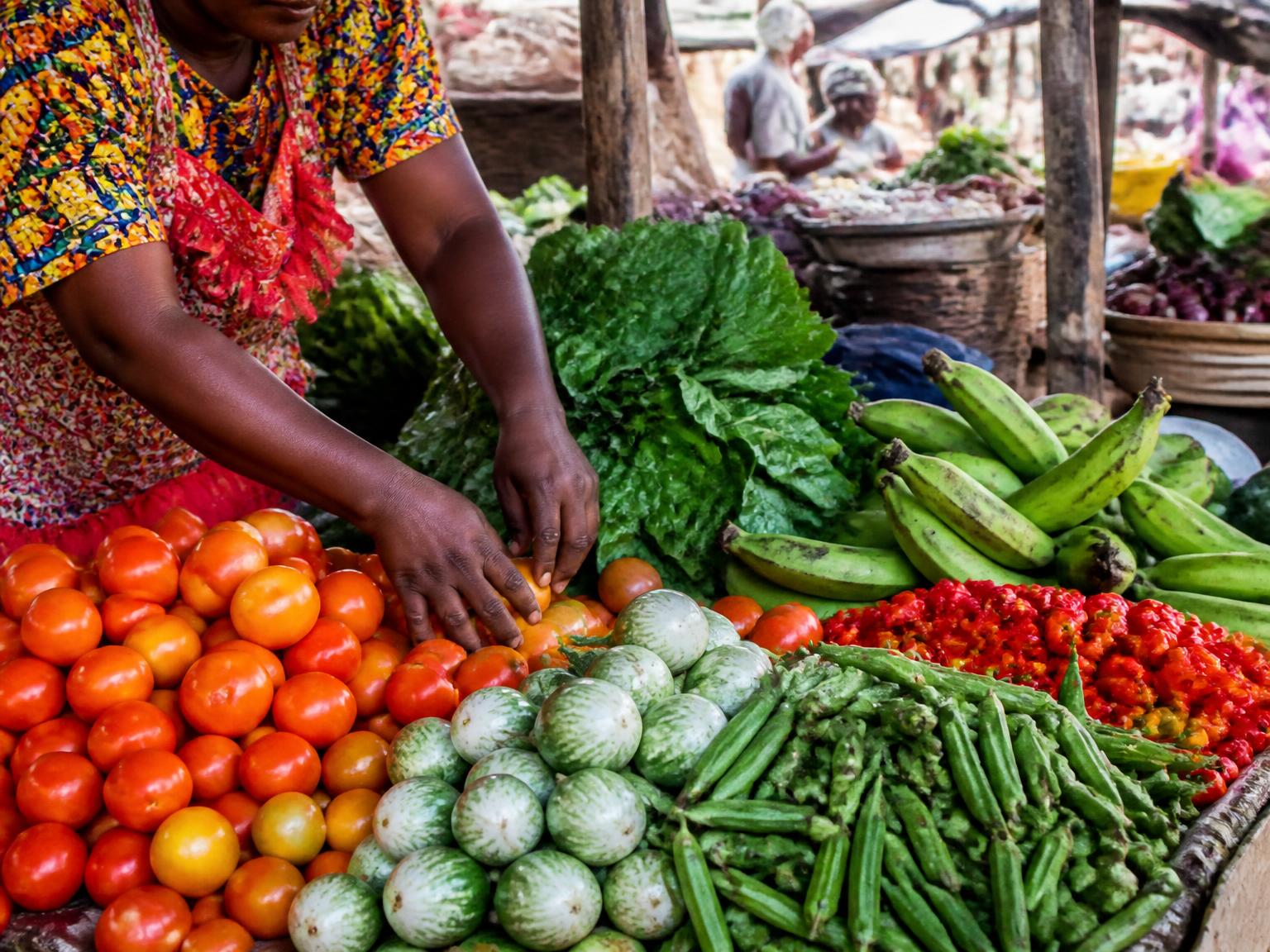 A market vendor arranging tomatoes, garden eggs, okra, plantain and leafy greens on a stall